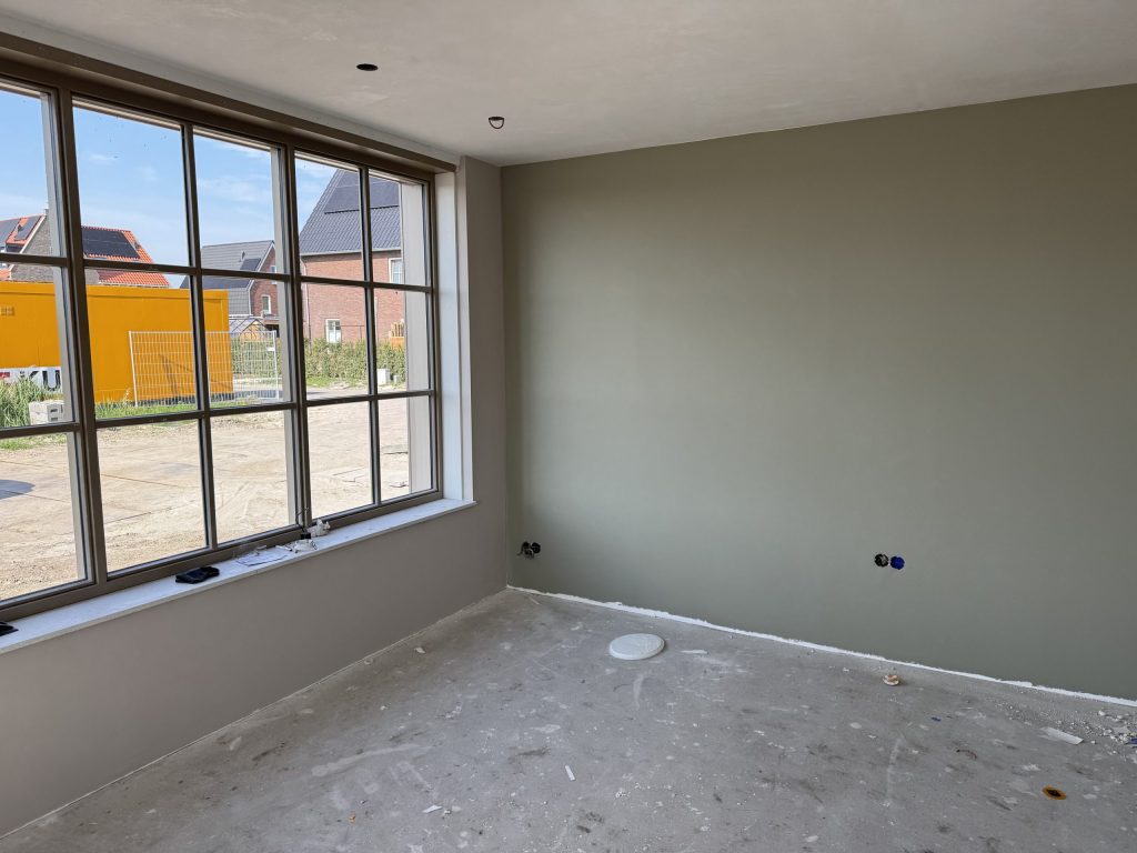 Kamer in een nieuw huis met een wand in grijsgroen en een groot raam met zicht op de bouwplaats
